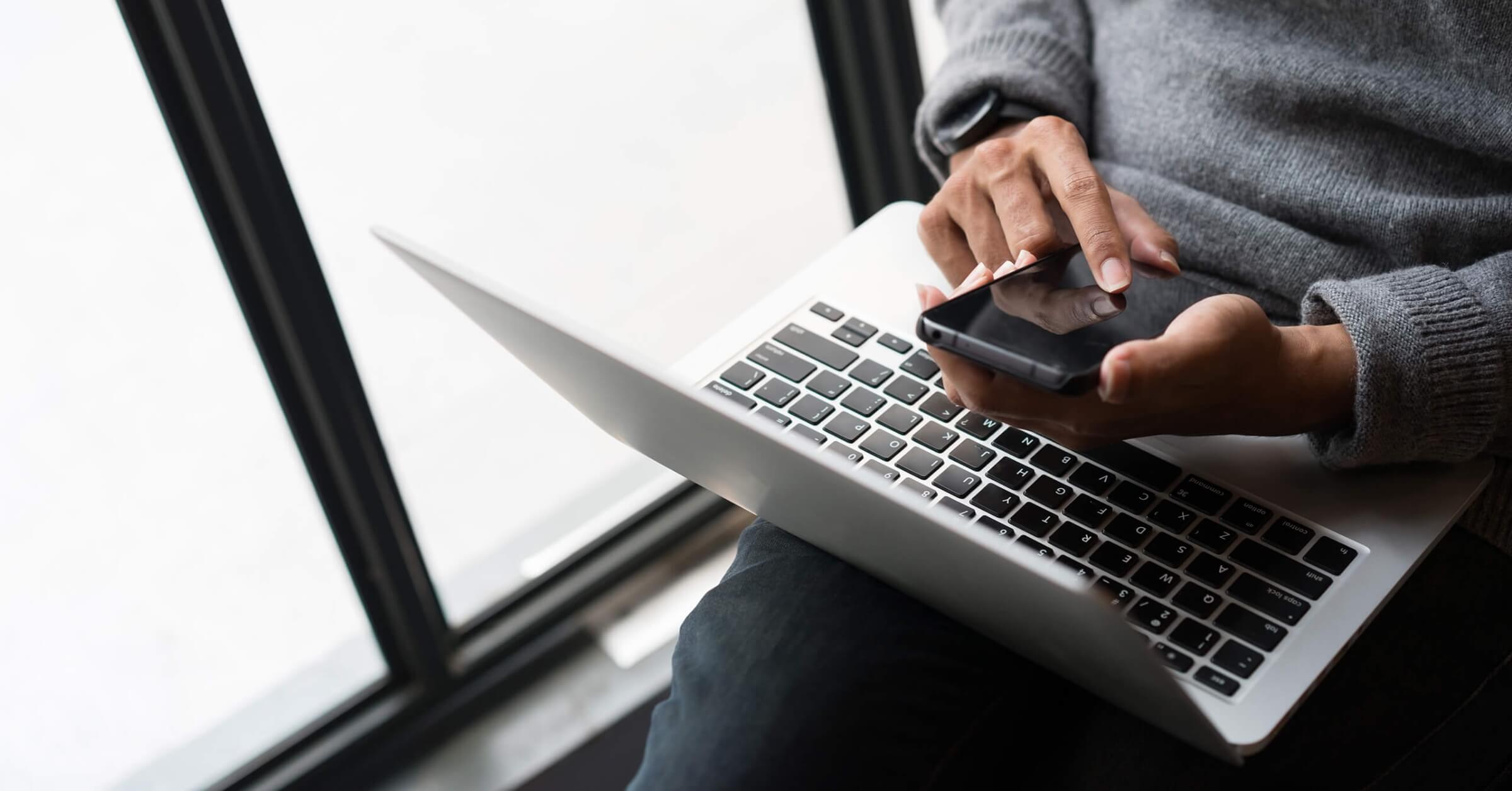 Mann med grå genser sitter ved vinduet med laptop på fanget og telefon i hånden